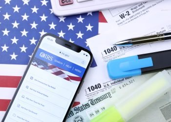 KHARKOV, UKRAINE - MARCH 5, 2021: Official IRS website on iPhone 12 pro screen with 1040 and w-2 paper tax forms on United States flag